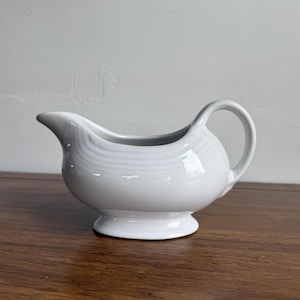May include: A white ceramic gravy boat with a rounded body, a spout, and a handle. The gravy boat sits on a small, circular base. The surface has a subtle, textured pattern. The background is a light gray wall.
