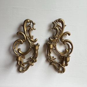May include: A pair of gold-coloured wall sconces featuring ornate scrollwork and floral detailing. Each sconce includes two cylindrical candle holders integrated into the curved, decorative frame, designed for mounting on a vertical surface.