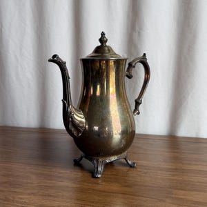 Puede incluir: Una cafetera vintage plateada con un pico curvo y un asa ornamentada. La cafetera tiene una tapa con un adorno decorativo y descansa sobre tres pequeños pies. La superficie metálica refleja la luz.