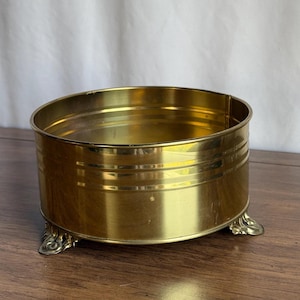 May include: A round, brass-coloured container with a decorative design. The container has a cylindrical shape with three small, ornate feet. The exterior features horizontal etched lines. The container is sitting on a wooden surface.