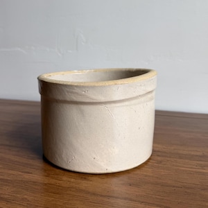 May include: A cylindrical ceramic crock with a light beige, matte finish. The vessel features a thick, slightly raised rim and a smooth, unadorned exterior surface. The stoneware container has a wide, open top and a flat base, showing subtle texture.