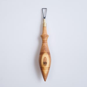 May include: A wood carving tool with a light brown, teardrop-shaped handle. The handle has a brass ferrule and a black metal blade. The initials "RD" are engraved on the handle. The tool is designed for detailed woodworking and carving.