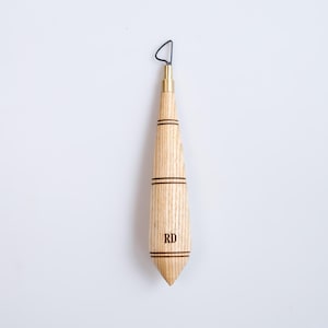 Op de afbeelding: Een houten klei-beeldhouwgereedschap met een metalen lus aan de bovenkant. De handgreep is lichtgekleurd hout met twee donkere banden en de initialen "RD". De tool heeft een messing fitting en een puntig uiteinde.