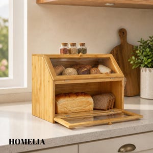 May include: A wooden bread bin with a clear glass door and top, showcasing different types of bread. The bin has two compartments and is labelled "HOMELIA". Three small glass jars with cork lids are placed on top of the bread bin.