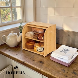 May include: A natural bamboo bread bin with three shelves, holding assorted loaves and pastries. A white teapot and a small plant are on the counter. The word "HOMELIA" is visible at the bottom.