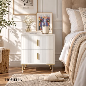 May include: A white two-drawer bedside table featuring a vertical ribbed texture on the drawer fronts, slim gold vertical handles, and four gold-toned metal legs. The bedside table is positioned in a bedroom setting with a book and mug on top.