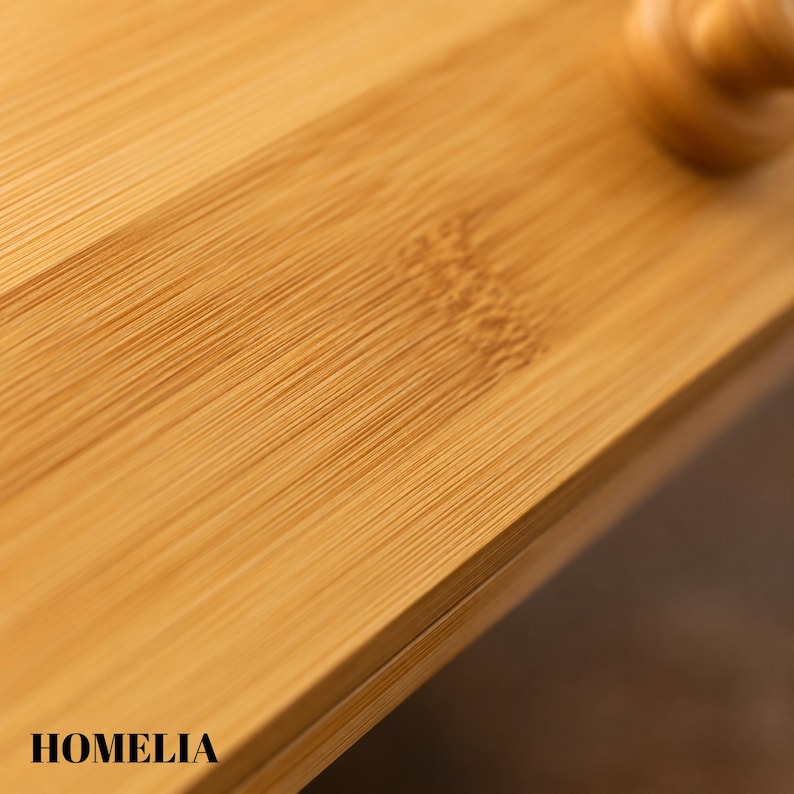 May include: Close-up of a bamboo cutting board with visible wood grain. The board is a light brown colour and has the word "HOMELIA" printed on the bottom left corner. The surface is smooth and appears to be well-crafted.