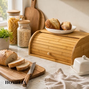 May include: A bamboo bread bin with a roll-top lid, a wooden chopping board with sliced bread, and a butter dish. Jars of pasta and oats are visible in the background. A bread knife rests on the chopping board. The word "HOMELIA" is printed on the chopping board.