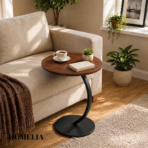 May include: A circular, dark brown side table with a black, curved metal leg and base. A cup of coffee, a small plant, and a book rest on the table. The table is 51 cm in diameter.