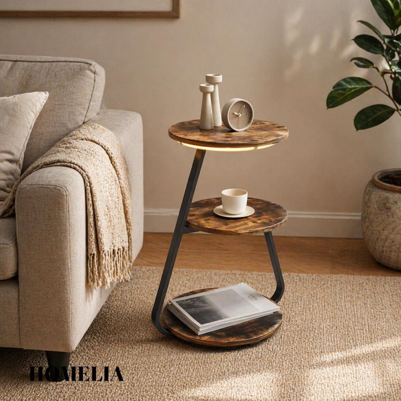 May include: A three-tier side table with a black metal frame and round, dark wood-grain shelves. The top shelf features an integrated LED light strip, two small decorative pillars, and a round clock. A white cup and a book are on the lower shelves.