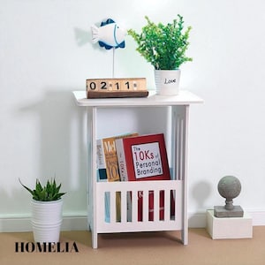 May include: A white wooden side table with a slatted magazine rack. The table holds books, a small potted plant, and a decorative fish. A wooden block calendar displays the date. The word "HOMELIA" is at the bottom.