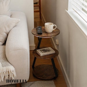 May include: A three-tiered round side table with a dark wood grain finish and black metal frame. The top tier holds a white ceramic mug and a black remote control, while the middle tier displays a book titled Didion. The bottom tier is empty.