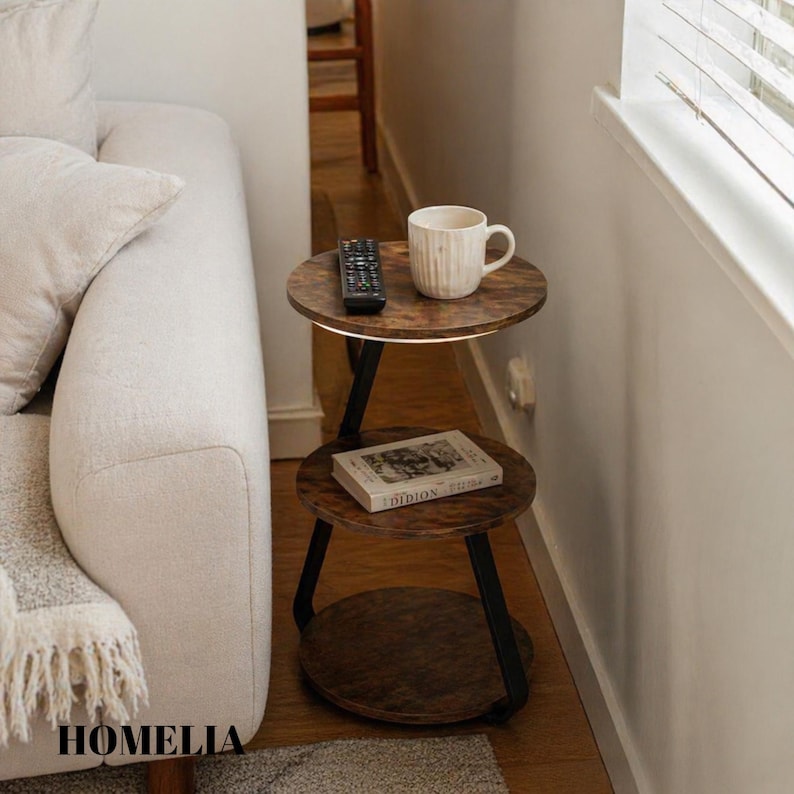 May include: A three-tiered round side table with a dark wood grain finish and black metal frame. The top tier holds a white ceramic mug and a black remote control, while the middle tier displays a book titled Didion. The bottom tier is empty.