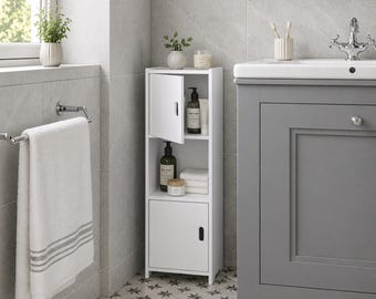 May include: A white bathroom storage cabinet with a partially open door, holding toiletries and towels. The cabinet is next to a gray vanity and a towel rack with a white towel. The floor has a star pattern. The word "HOMELIA" is at the bottom.