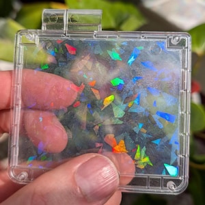 Könnte beinhalten: Ein klarer, rechteckiger, klappbarer Kasten, gefüllt mit schillernden, farbenfrohen, geometrischen Formen. Der Kasten wird von einer Hand gehalten, mit verschwommenem grünem Laub im Hintergrund.