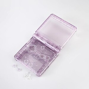 May include: A clear purple plastic shell for a handheld game console. The shell is open and shows the buttons and screen area. There are two small clear plastic pieces next to the shell.
