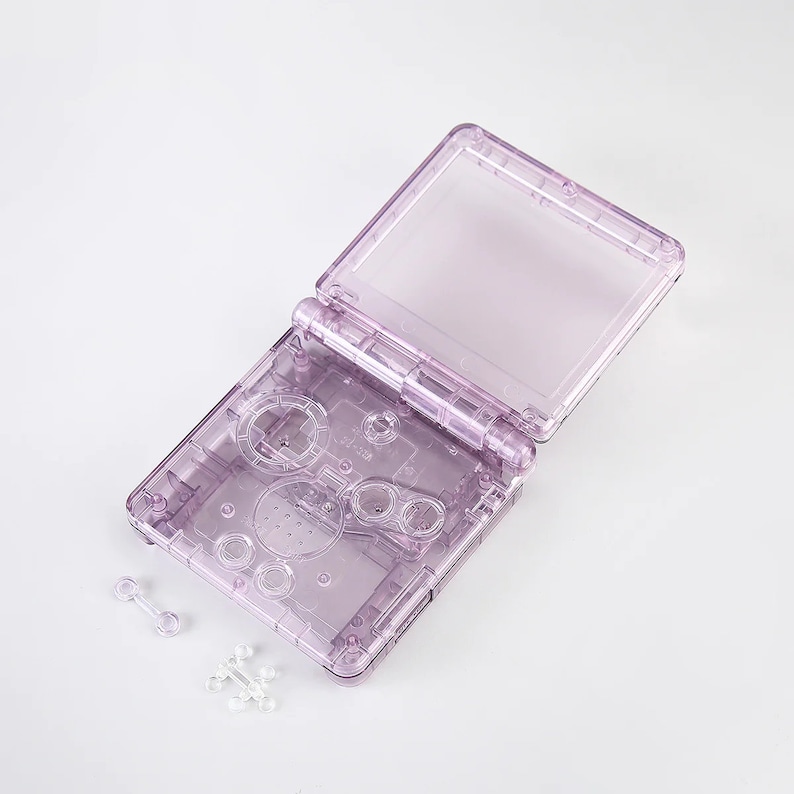 May include: A clear purple plastic shell for a handheld game console. The shell is open and shows the buttons and screen area. There are two small clear plastic pieces next to the shell.