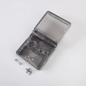 May include: A clear plastic shell for a handheld video game console. The shell is open and the buttons and screen are visible. There are two small metal pieces next to the shell.