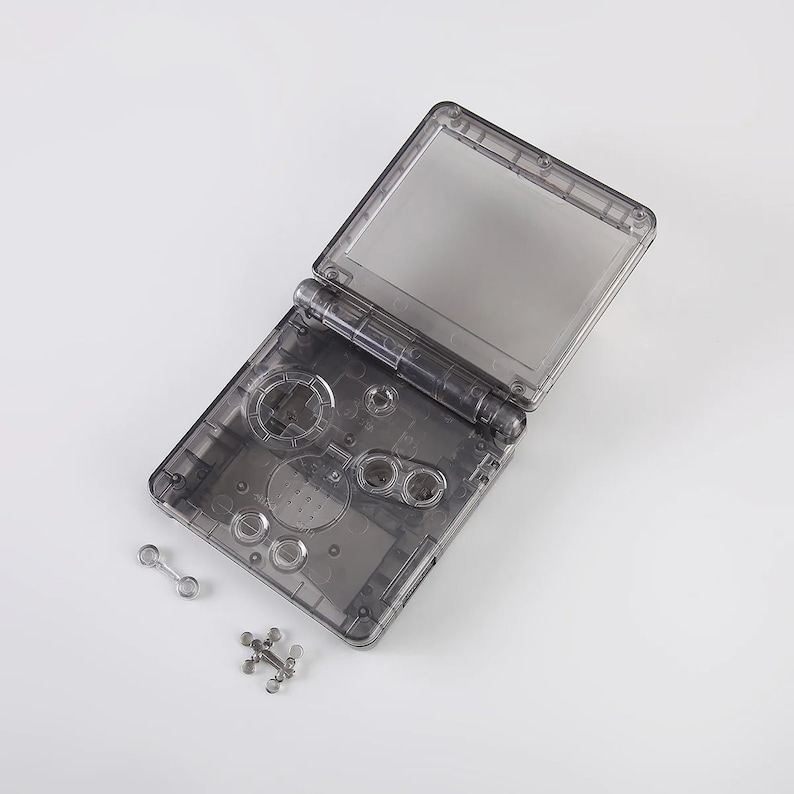 May include: A clear plastic shell for a handheld video game console. The shell is open and the buttons and screen are visible. There are two small metal pieces next to the shell.