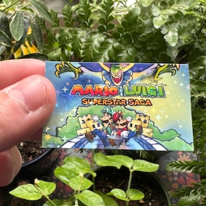 May include: A holographic card with a colorful image of Mario and Luigi from the video game "Mario & Luigi: Superstar Saga". The card has a blue background with stars and a white border.