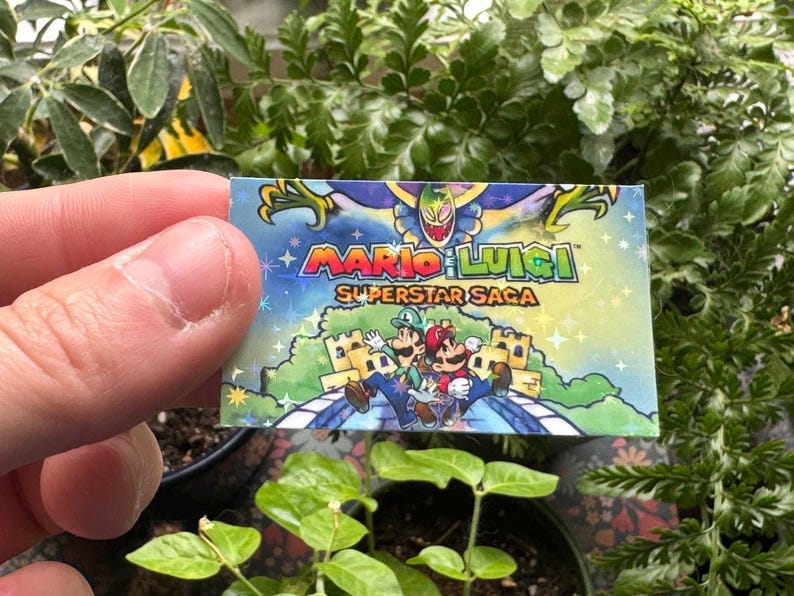 May include: A holographic card with a colorful image of Mario and Luigi from the video game "Mario & Luigi: Superstar Saga". The card has a blue background with stars and a white border.