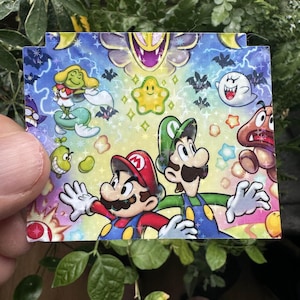 May include: A colorful card featuring Mario and Luigi from the Super Mario Bros. video game series. The card has a blue background with stars, bats, and other characters from the game.