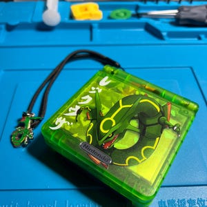 以下が含まれることがあります： 緑色の任天堂ゲームボーイアドバンスSP、レックウザのデザイン。ハンドヘルドコンソールは、黒いストラップとチャームが付いた緑色のケースです。レックウザのグラフィックは緑と黒です。