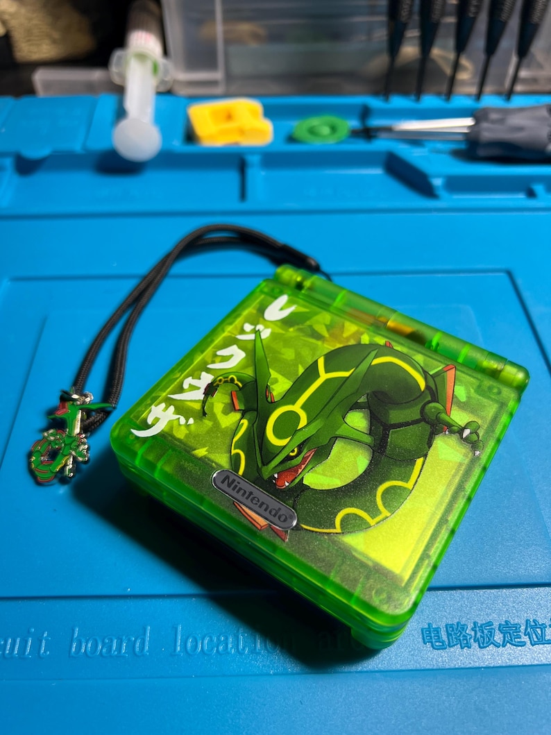 以下が含まれることがあります： 緑色の任天堂ゲームボーイアドバンスSP、レックウザのデザイン。ハンドヘルドコンソールは、黒いストラップとチャームが付いた緑色のケースです。レックウザのグラフィックは緑と黒です。