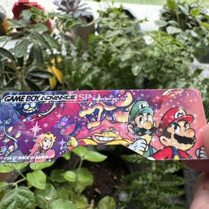May include: A close-up of a Game Boy Advance SP screen protector featuring a colorful design with Mario, Luigi, and other characters. The text "GAME BOY ADVANCE SP MODEL NO. AGS-101" is printed on the protector.