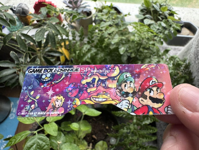 May include: A close-up of a Game Boy Advance SP screen protector featuring a colorful design with Mario, Luigi, and other characters. The text "GAME BOY ADVANCE SP MODEL NO. AGS-101" is printed on the protector.