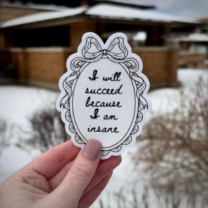 May include: A white sticker with a black border and the text "I will succeed because I am insane" in a decorative oval frame. The sticker has a bow at the top and is held by a hand.