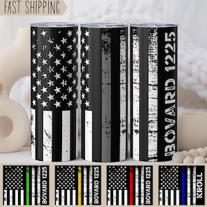 May include: Three stainless steel tumblers with a distressed American flag design. The tumblers feature a thin black line, a thin gray line, and a thin blue line, respectively. The tumbler on the right has the text "Boward 1225" printed on it.