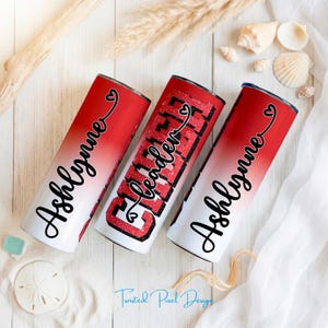 May include: Three red and white ombre tumblers with black text. Two tumblers have the name "Ashlynne" in a cursive font, and one says "Cheer Leader" in a bold, glittery font. Each tumbler has a heart design. The tumblers are on a white wooden surface.