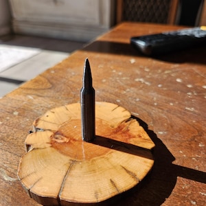 Könnte beinhalten: Eine dunkelgraue Patrone steht aufrecht auf einem kleinen, runden Holzsockel. Der Sockel besteht aus einem Querschnitt eines Astes, der die natürliche Holzmaserung und Farbvariationen zeigt. Die Patrone ist auf dem Holzsockel zentriert.