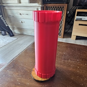 May include: A tall, cylindrical red container with a smooth body and textured, ribbed bands at the top and bottom edges. The object stands upright on a small, circular wooden base with visible natural bark edges and a light brown wood grain surface.