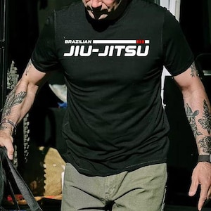 Puede incluir: Camiseta deportiva negra con las palabras "BRAZILIAN JIU-JITSU" en letras blancas. La camiseta tiene una raya roja encima del texto. La persona lleva pantalones verde oliva.