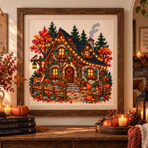 May include: A framed cross-stitch artwork depicting a cozy cottage surrounded by autumn foliage, a deer, and a squirrel. The scene features warm string lights, pumpkins, and a stone path. The artwork is displayed with fall-themed decor including books, candles, and a wooden sign with the text A Brighter Day One Stitch at a Time.