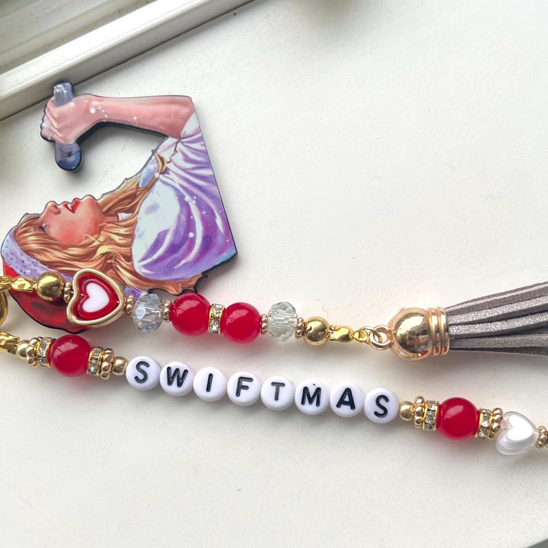 May include: A white beaded keychain with red beads, a gold charm, and a grey tassel. The keychain spells out "SWIFTMAS" in white letters. A small red heart charm with a white centre is attached to the keychain. A cartoon image of a woman singing into a microphone is attached to the keychain.