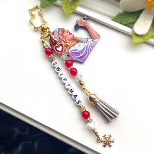 May include: A gold charm featuring a heart-shaped image of a woman singing into a microphone. The charm is attached to a beaded chain with red and white beads spelling out "SWIFTMAS". The chain ends with a grey tassel and a gold snowflake charm.