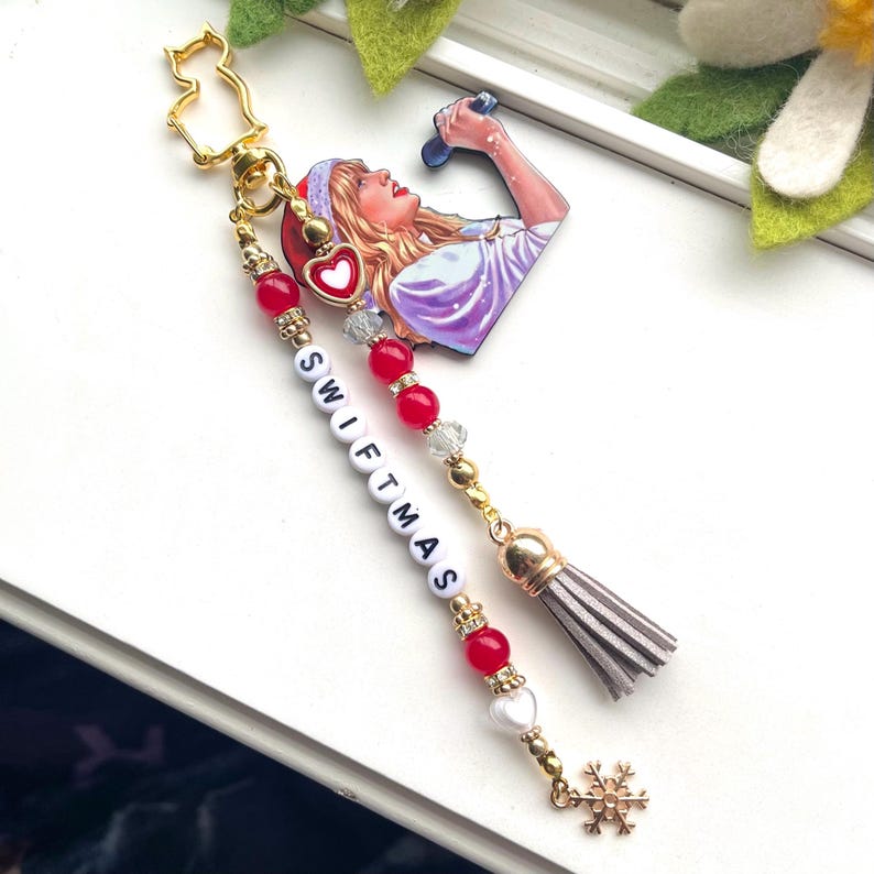 May include: A gold charm featuring a heart-shaped image of a woman singing into a microphone. The charm is attached to a beaded chain with red and white beads spelling out "SWIFTMAS". The chain ends with a grey tassel and a gold snowflake charm.