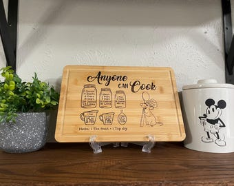 Puede incluir: Una tabla de cortar rectangular de bambú con el texto "Anyone Can Cook" y medidas de cocina. La tabla presenta un dibujo de un ratón de dibujos animados sosteniendo un globo. Un bote blanco de Mickey Mouse está a la derecha.