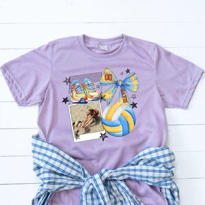 May include: Purple t-shirt with a volleyball graphic. The graphic features a volleyball, a bow with the word "PLAYER" on it, a pair of sneakers, and a photo of a volleyball player. The graphic is surrounded by stars.