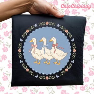 May include: Black square pillow featuring three cartoon geese with pink, blue, and pink bows, inside a blue circle with a floral border. The pillow is held by a hand, and the text "ChipChipLady" is in the upper right corner.