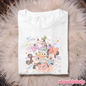 May include: White t-shirt featuring a colorful illustration of Disney characters Mickey Mouse, Minnie Mouse, Donald Duck, Daisy Duck, Goofy, and Pluto. The design includes the characters' names and decorative elements. The shirt is folded on a fluffy, white surface.
