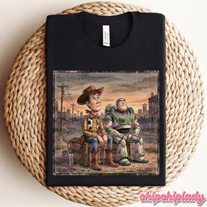 May include: Black t-shirt featuring a distressed graphic of Woody and Buzz Lightyear from Toy Story. Woody is in a cowboy outfit, and Buzz is in a spacesuit. The graphic is set against a cityscape backdrop.