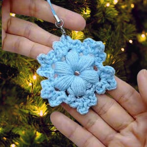 May include: A light blue, crocheted snowflake ornament with a central flower design. The snowflake is held by a blue string and a silver clasp. The ornament is displayed against a blurred background of a Christmas tree.