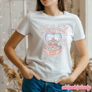 Może przedstawiać: Biała koszulka z kolorową ilustracją Disneylandu w pudełku w kształcie serca. Projekt zawiera zamek, fajerwerki i słowo "Disneyland" w fantazyjnej czcionce. Koszulka ma klasyczny okrągły dekolt.