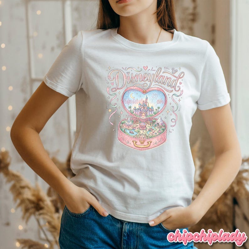 Może przedstawiać: Biała koszulka z kolorową ilustracją Disneylandu w pudełku w kształcie serca. Projekt zawiera zamek, fajerwerki i słowo "Disneyland" w fantazyjnej czcionce. Koszulka ma klasyczny okrągły dekolt.