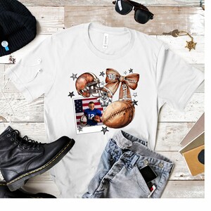 May include: White t-shirt with a football helmet, football, and a bow with the text "Davis" on it. A photo of a young boy wearing a football uniform is in the corner of the shirt.