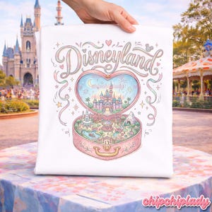 Może przedstawiać: Biała koszulka z ilustracją Disneylandu. Projekt przedstawia pudełko w kształcie serca z miniaturową sceną parku, napis "Disneyland" w ozdobnej czcionce i pastelowe elementy dekoracyjne. Koszulka jest trzymana przez dłoń.